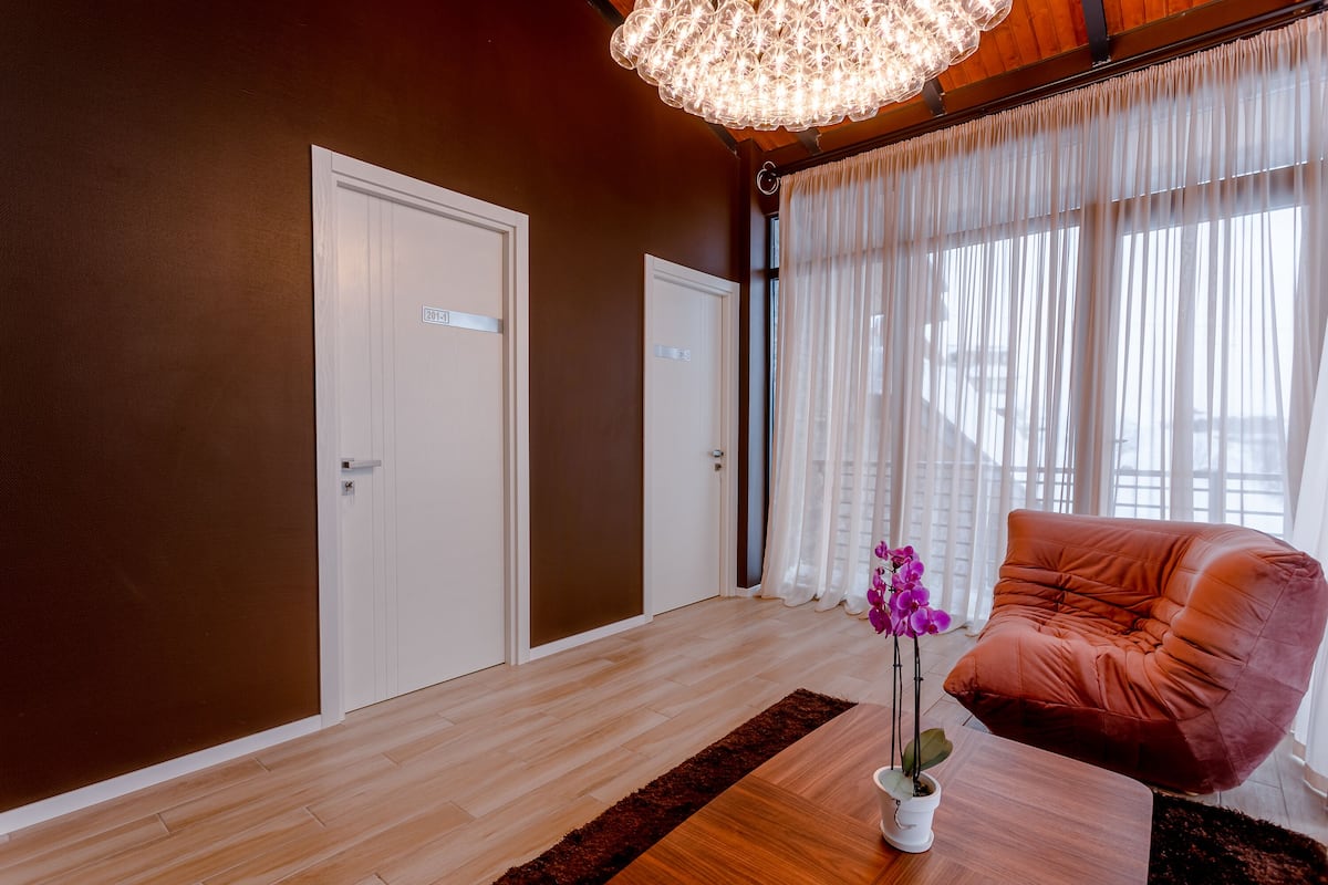 A room with a wooden floor, a large window with sheer curtains, a plush orange chair, and a white door with a sign.