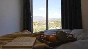 Een bed met een boek, sinaasappels en een dienblad, een raam met uitzicht op een landschap, en donkere gordijnen.
