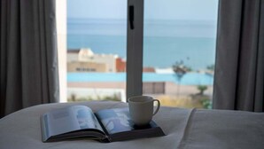 A bed with an open book and a mug, a window with a view of a pool and buildings.