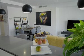 A modern living room with a dining area, a flat-screen TV, and a fruit display on the table.
