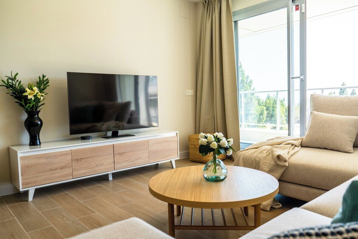 A modern living room with a flat-screen TV, a wooden coffee table, a beige sofa, and a large window with curtains.