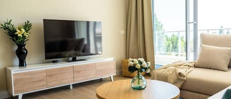 A modern living room with a flat-screen TV, a wooden coffee table, a beige sofa, and a large window with curtains.