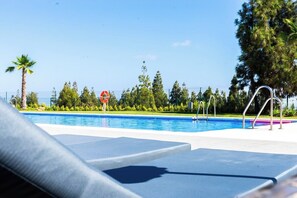 Una piscina con sillas de descanso, quiosco de salvavidas y un área cercada con árboles, incluyendo una palmera.