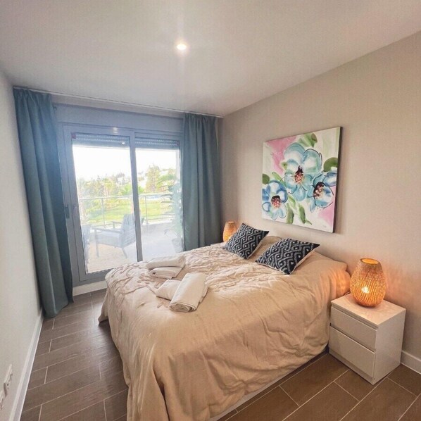 Un dormitorio con una cama grande, una puerta corrediza al balcón, un cuadro en la pared y una lámpara de noche.