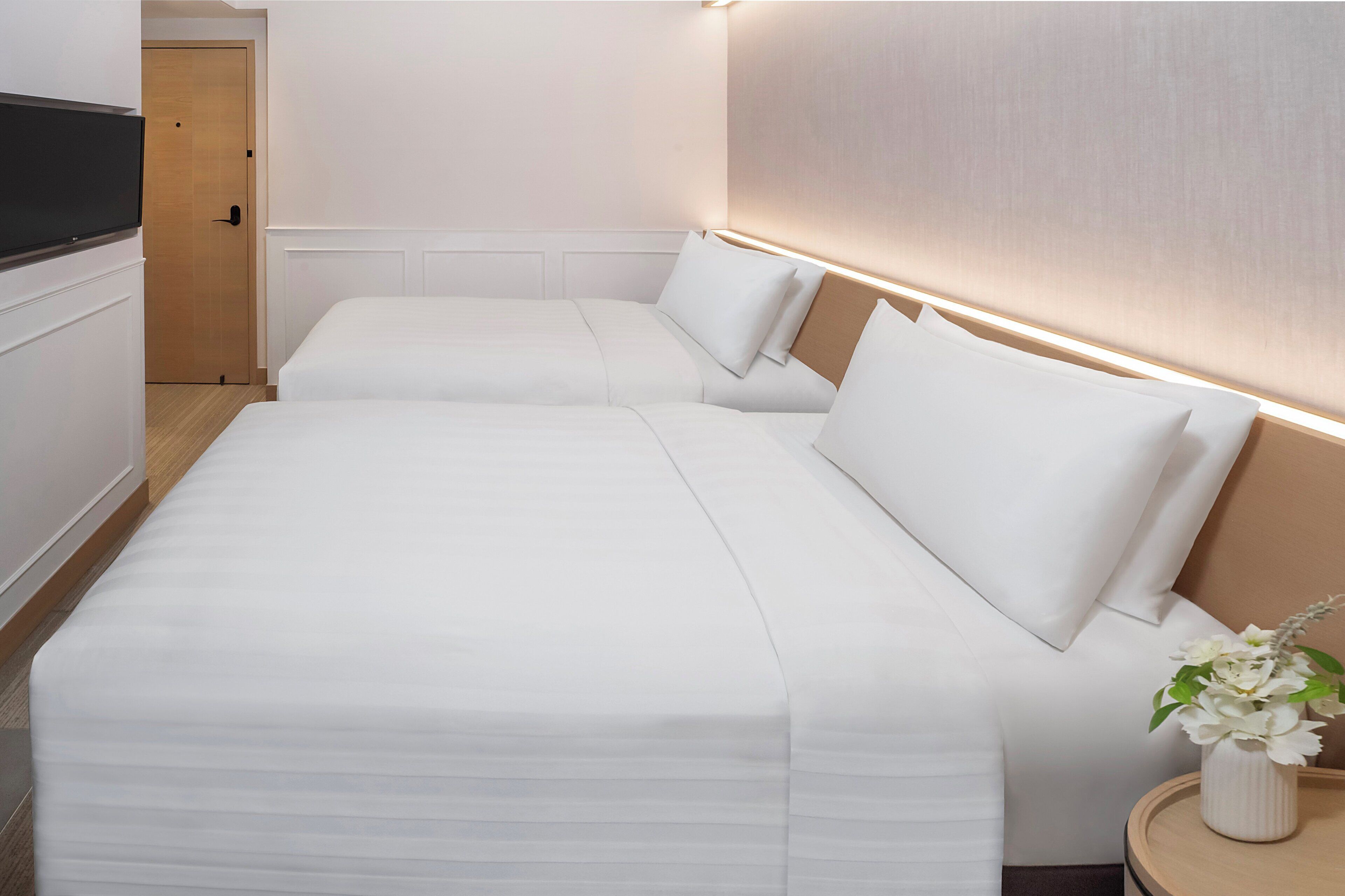 A hotel room with two beds, a wooden door, and a bedside table with a vase of flowers.