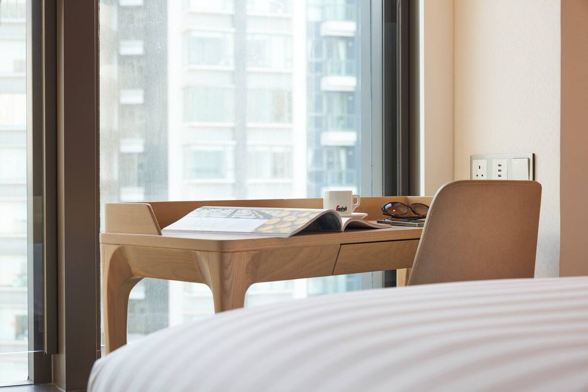 A hotel room with a large window, a wooden desk with a laptop, a cup, and a pair of glasses.