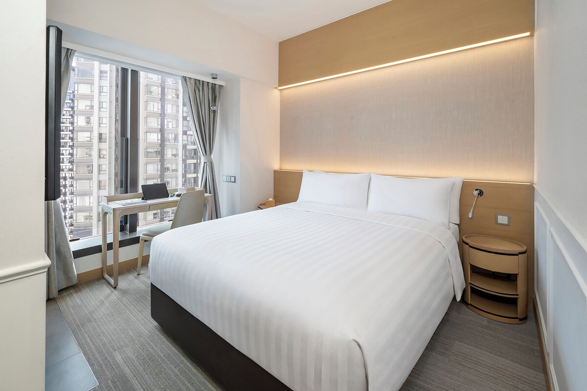 A hotel room with a large bed, a desk with a chair, a nightstand, and a window with curtains.