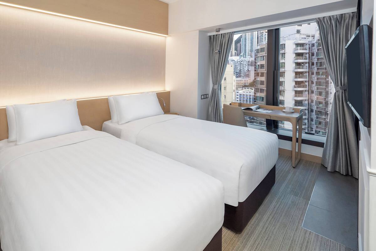 A hotel room with two beds, a small table, and a view of a cityscape through large windows.