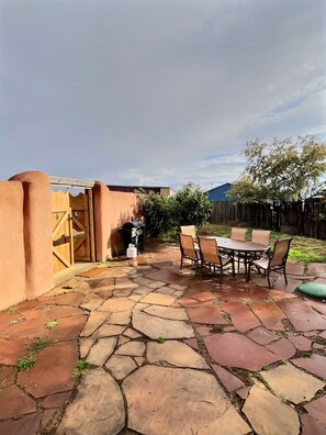 Eine Terrasse mit Steinbelag, einem Holztisch mit Stühlen, einem Grill und einem hölzernen Tor.