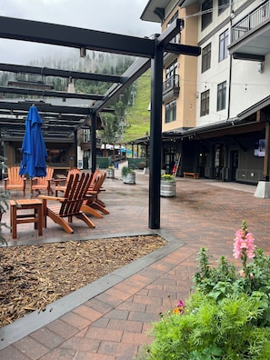 Relax in this tranquil outdoor seating area, featuring comfortable chairs and tables under a covered roof, surrounded by lush greenery and a charming garden.