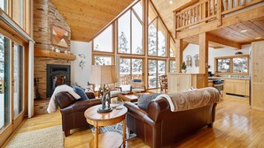 A spacious living room with a fireplace, leather sofas, a dining area, and large windows offering views of a snowy landscape.