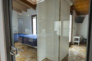 Une chambre avec une porte en verre donnant sur une salle de bain, un lit recouvert d’une couverture bleue et une petite table blanche.