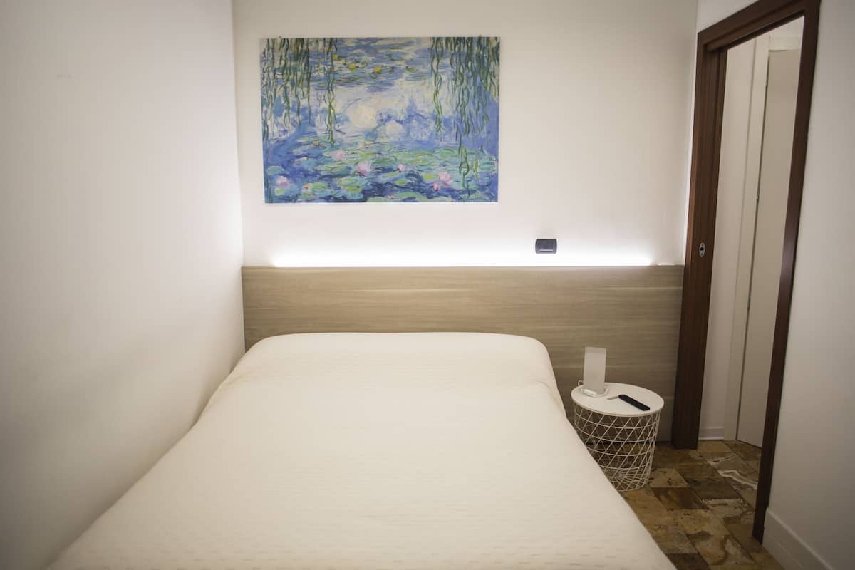 Un lit bien fait, avec une tête de lit, une petite table de chevet avec une lampe et un tableau représentant un étang, accroché au mur.