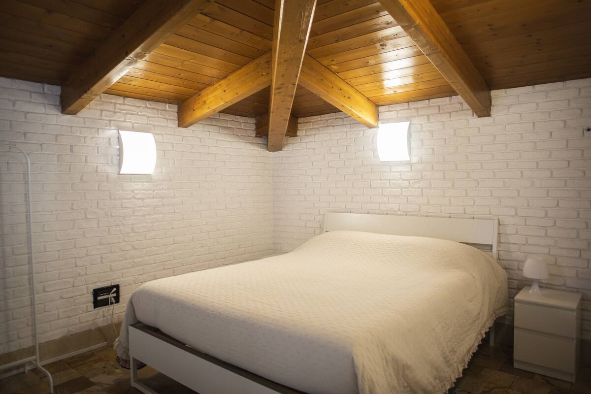 Une chambre avec un mur en briques blanches, un plafond en bois, un lit blanc et une table de chevet avec une lampe.