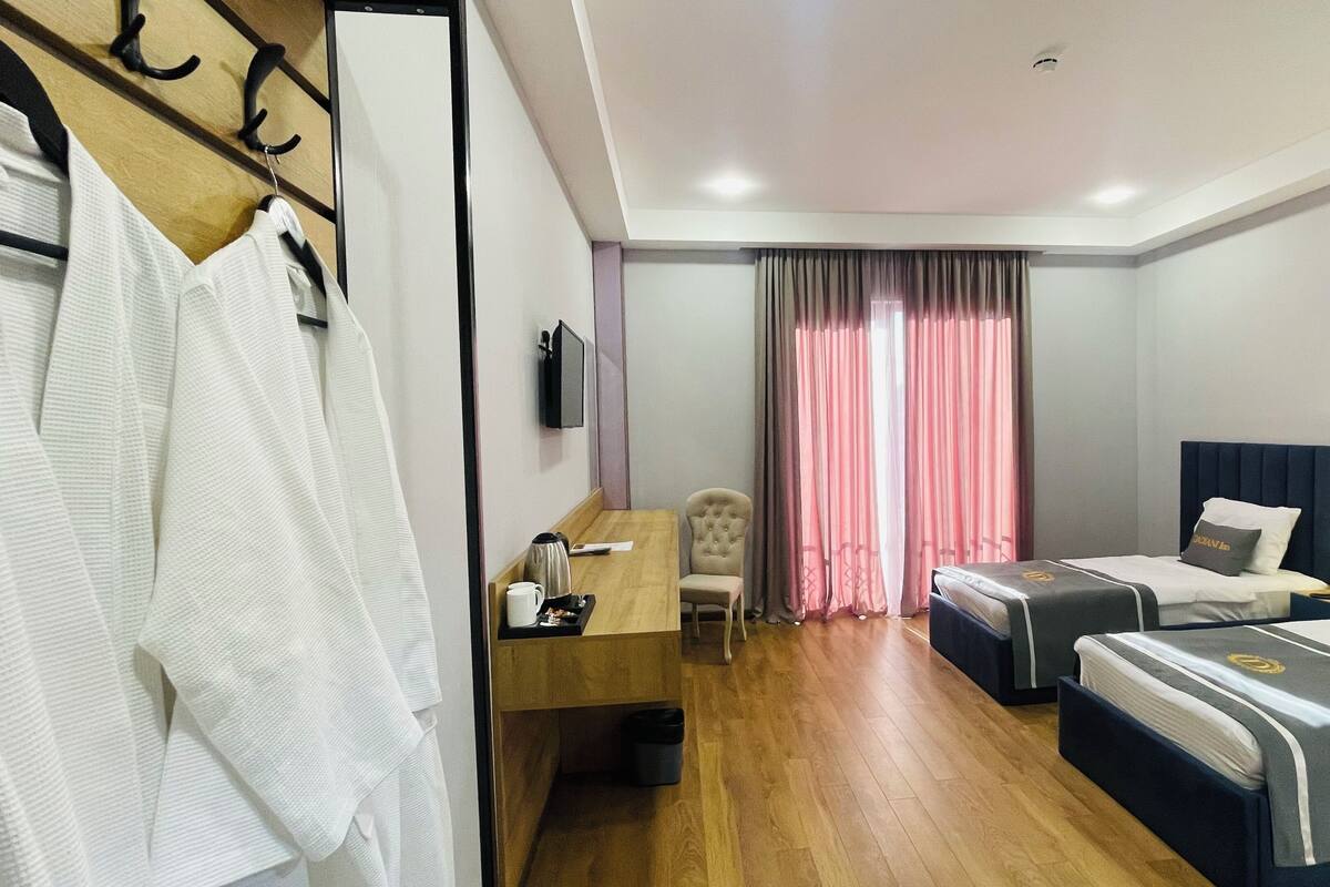A hotel room with a wooden floor, a desk with a chair, a bed with a headboard, and a wardrobe with hanging clothes.