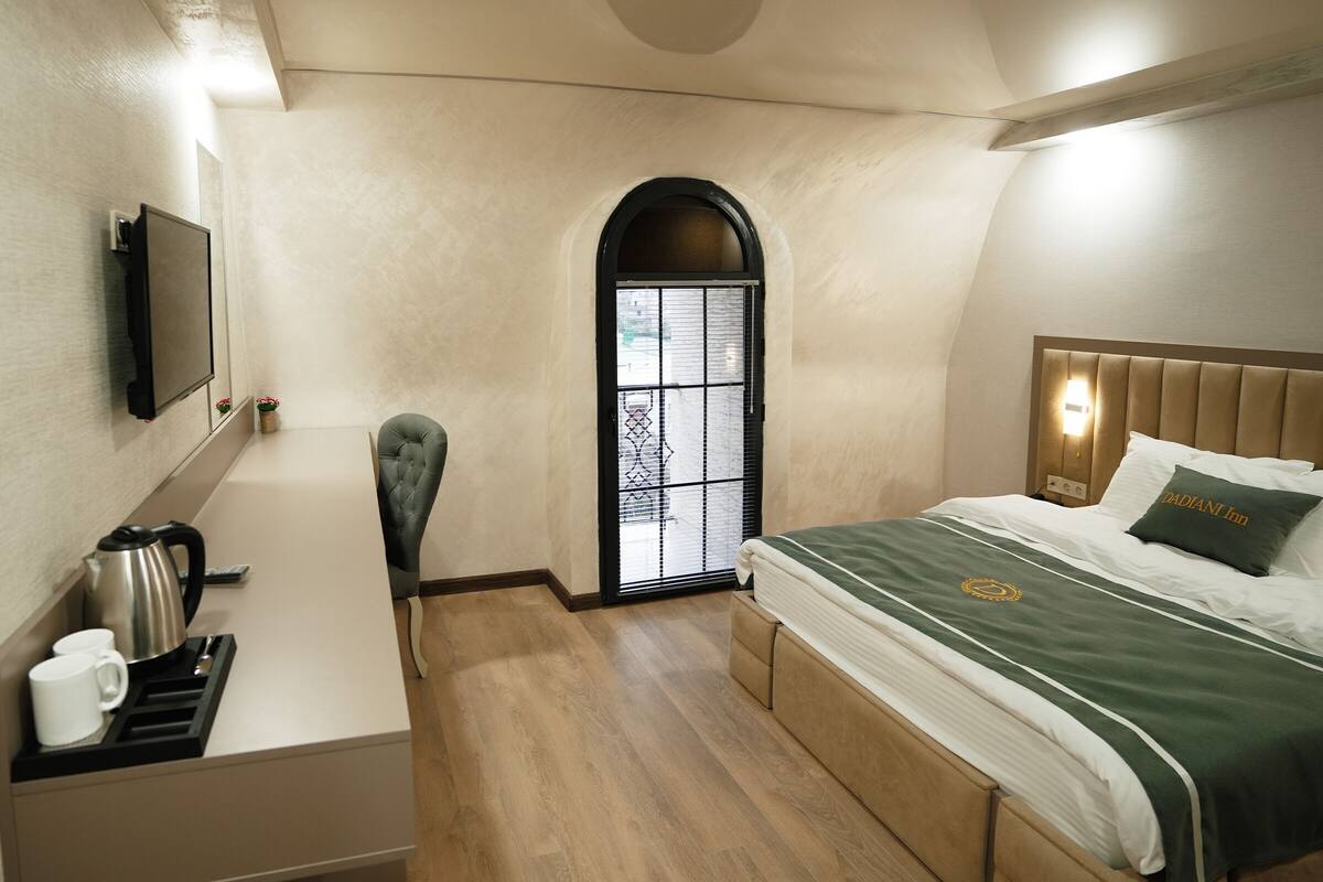 A hotel room with a bed, a desk with a kettle, a chair, and a television on the wall.
