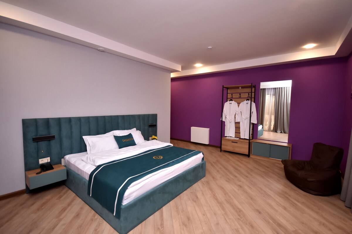 A modern hotel room with a large bed, a bedside table with a phone, a wardrobe with hanging clothes, a mirror, and a brown leather chair.