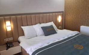 A hotel room with a padded headboard, a bedside phone, and a bed with white bedding and a grey pillow with "DADIANI" embroidered.