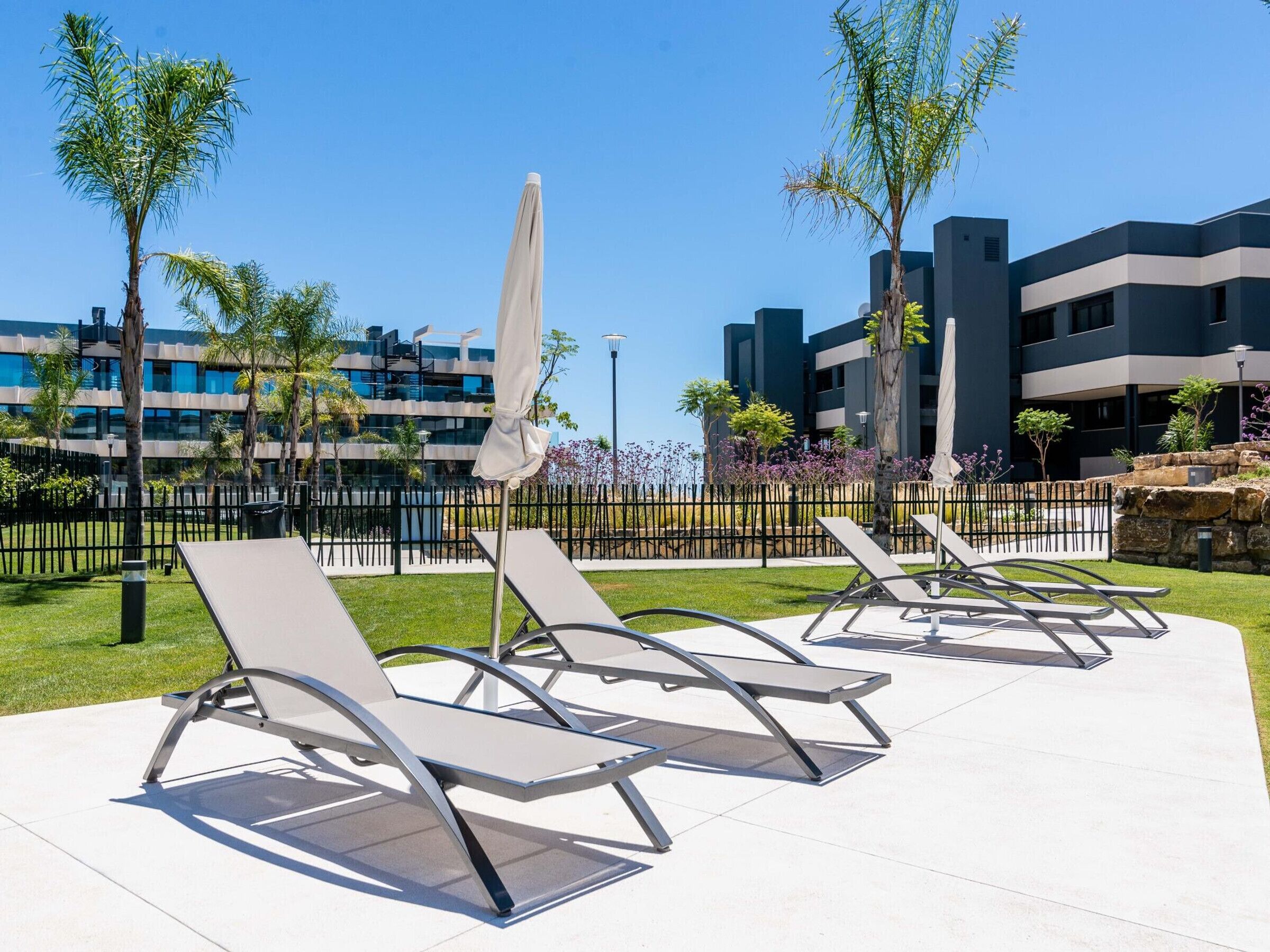 A modern outdoor seating area with sun loungers and umbrellas, surrounded by a well-maintained lawn and palm trees, with contemporary buildings in the background.
