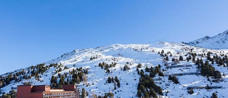 A hotel with a red facade and multiple floors, situated on a snow-covered mountain with pine trees.