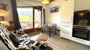 A cozy living room with a cow-print sofa, a dining area with a table and chairs, a kitchen with a refrigerator and oven, and a large window offering a view of the outdoors. - 2-room apartment in Praz Sur Arly, parking, close to slopes, for 5 people (Praz-sur-Arly)