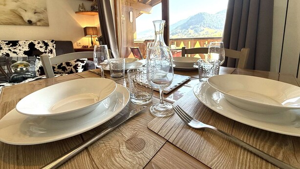 A dining table set with plates, glasses, and cutlery, with a view of mountains through large windows. - 2-room apartment in Praz Sur Arly, parking, close to slopes, for 5 people (Praz-sur-Arly)