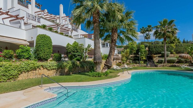 Una piscina con agua cristalina azul, rodeada de palmeras y un edificio blanco con balcones.