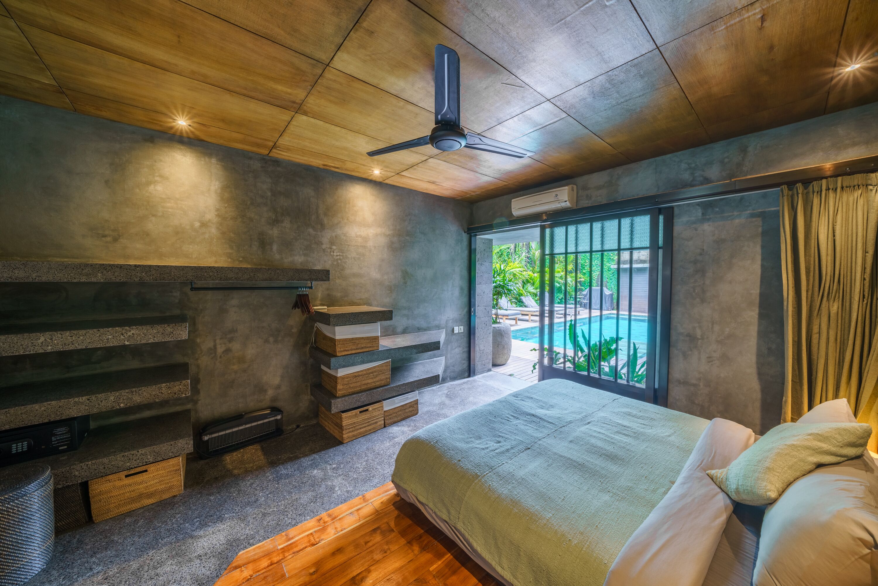 Une chambre à coucher comprenant un lit, un ventilateur de plafond, un banc intégré avec des tiroirs, une fenêtre donnant sur un espace verdoyant et un appareil de climatisation.