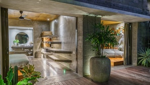 A modern bathroom with a large mirror, a sink, and a shower area. There is a wooden bench and a large potted plant in the room. - Love Supreme Villa by Betterplace (Canggu)