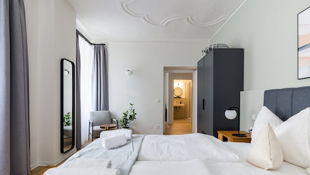 Une chambre d’hôtel moderne avec un grand lit, une table de chevet, une chaise, une armoire et une salle de bain visible à travers une porte ouverte.