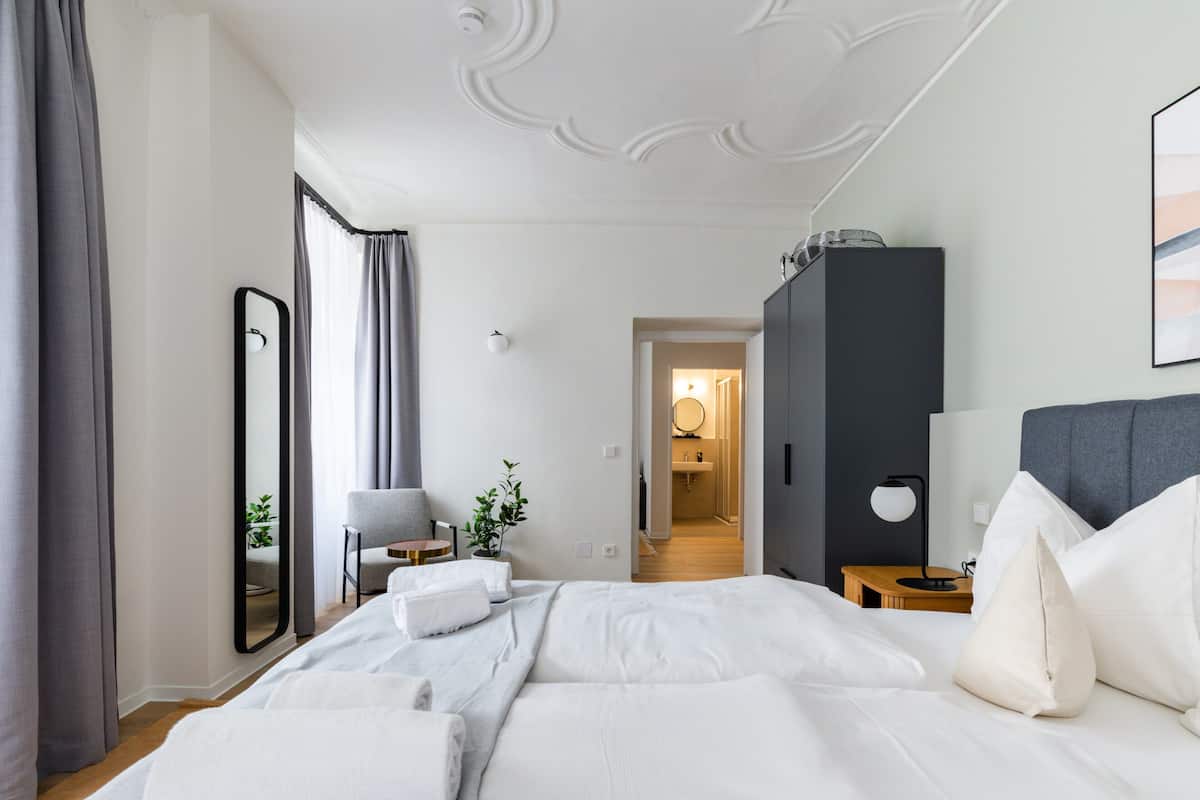 Quarto de hotel moderno com uma cama grande, mesa de cabeceira, poltrona, guarda-roupa e banheiro visível por uma porta aberta.