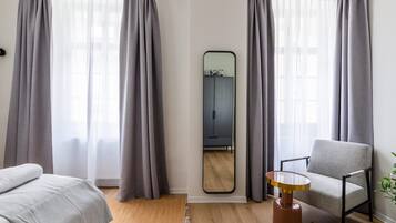 Una habitación de hotel moderna con cortinas grises, suelo de madera, sofá gris, una mesa redonda y un espejo.