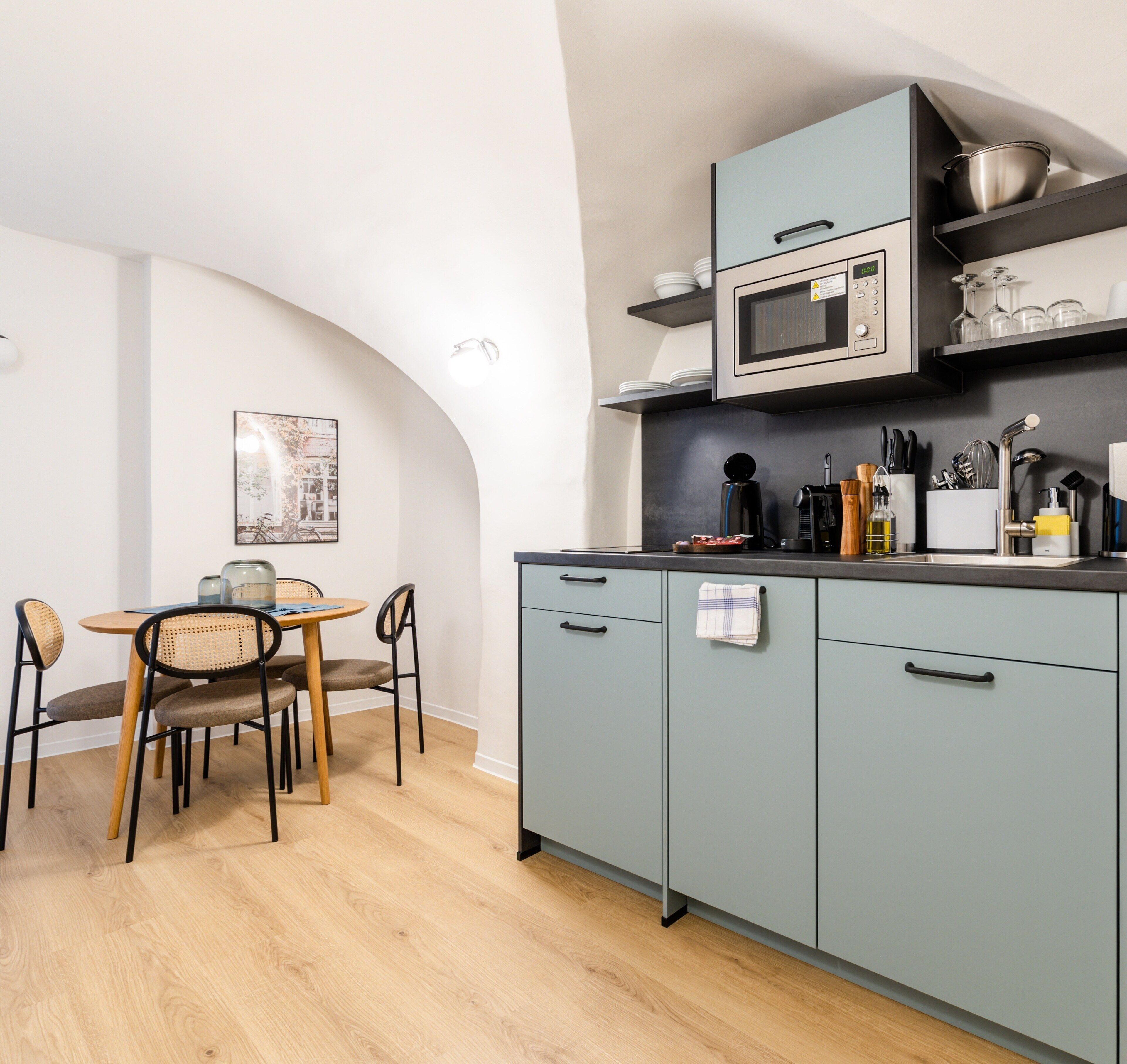 A modern kitchen with grey cabinets, a microwave, and a coffee machine. There is a dining table with chairs and a framed picture on the wall.
