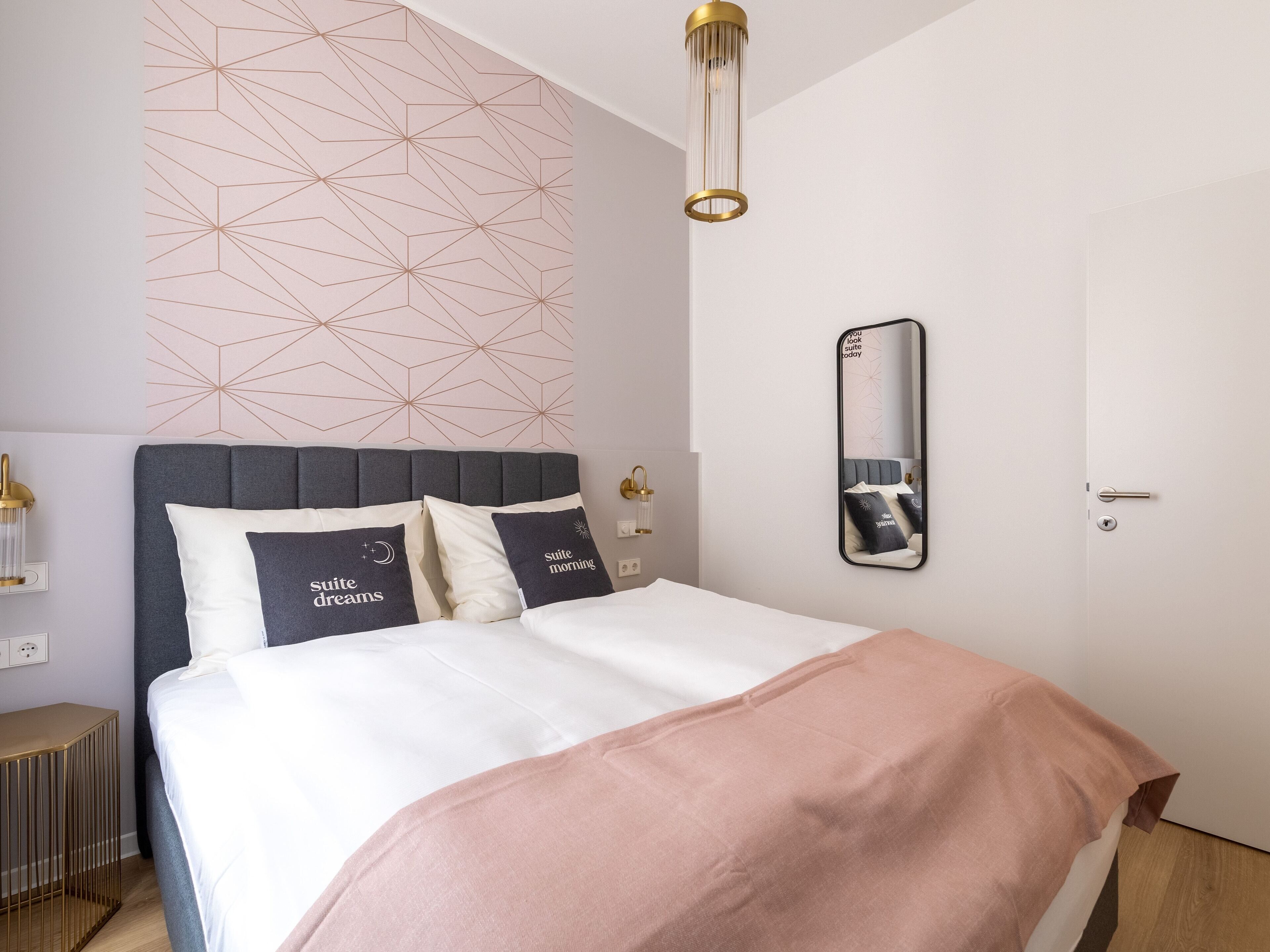A modern bedroom with a geometric-patterned wall, a large bed with a grey headboard, and a bedside table with a lamp.