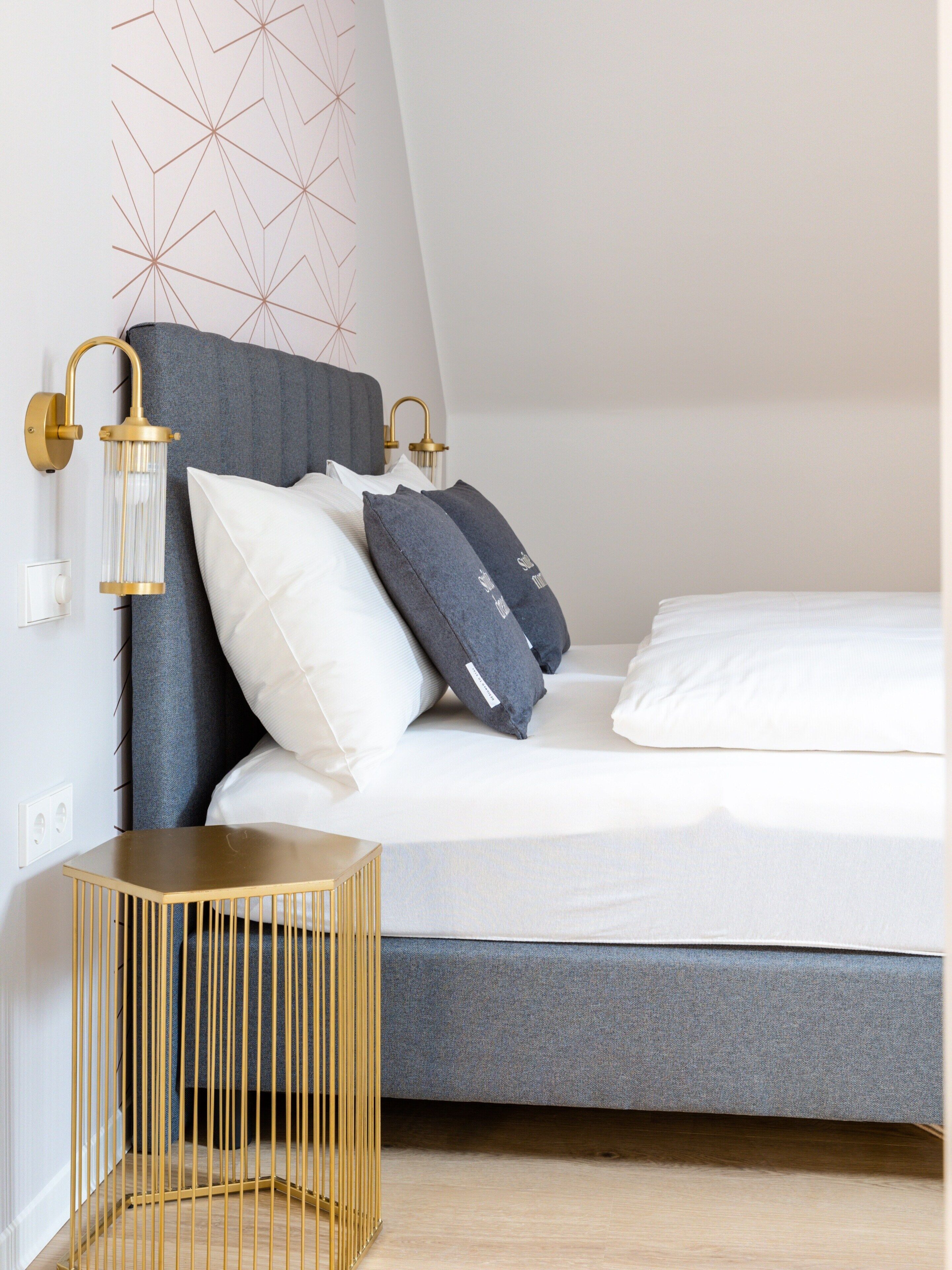 A modern bedroom with a grey headboard, white and dark grey pillows, a bedside table, and a wall-mounted light fixture.