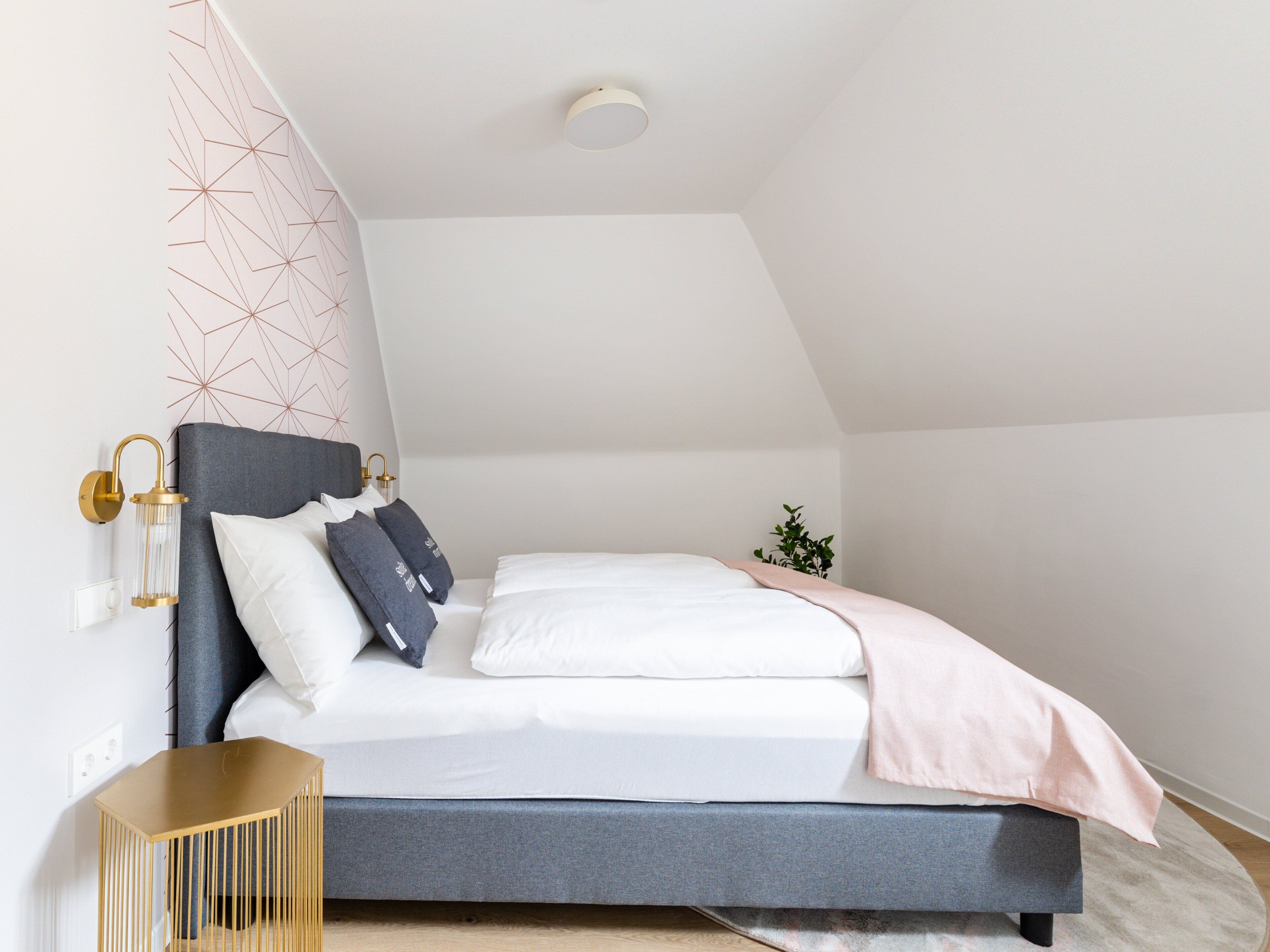 Un dormitorio moderno con techo inclinado, una cama con ropa de cama blanca y gris, una mesita de noche pequeña y un panel decorativo en la pared.