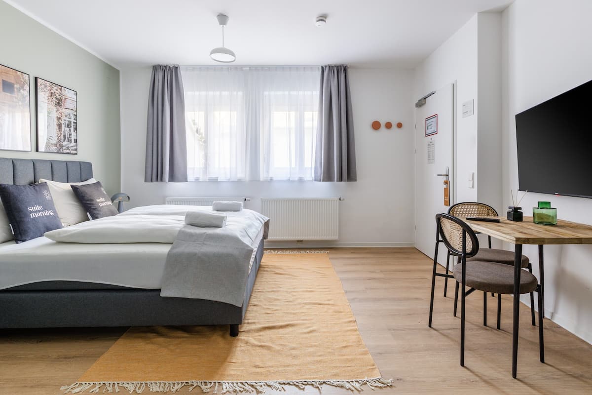 Quarto com cama, janela com cortinas, radiador, escrivaninha com cadeira e televisão de tela plana na parede.