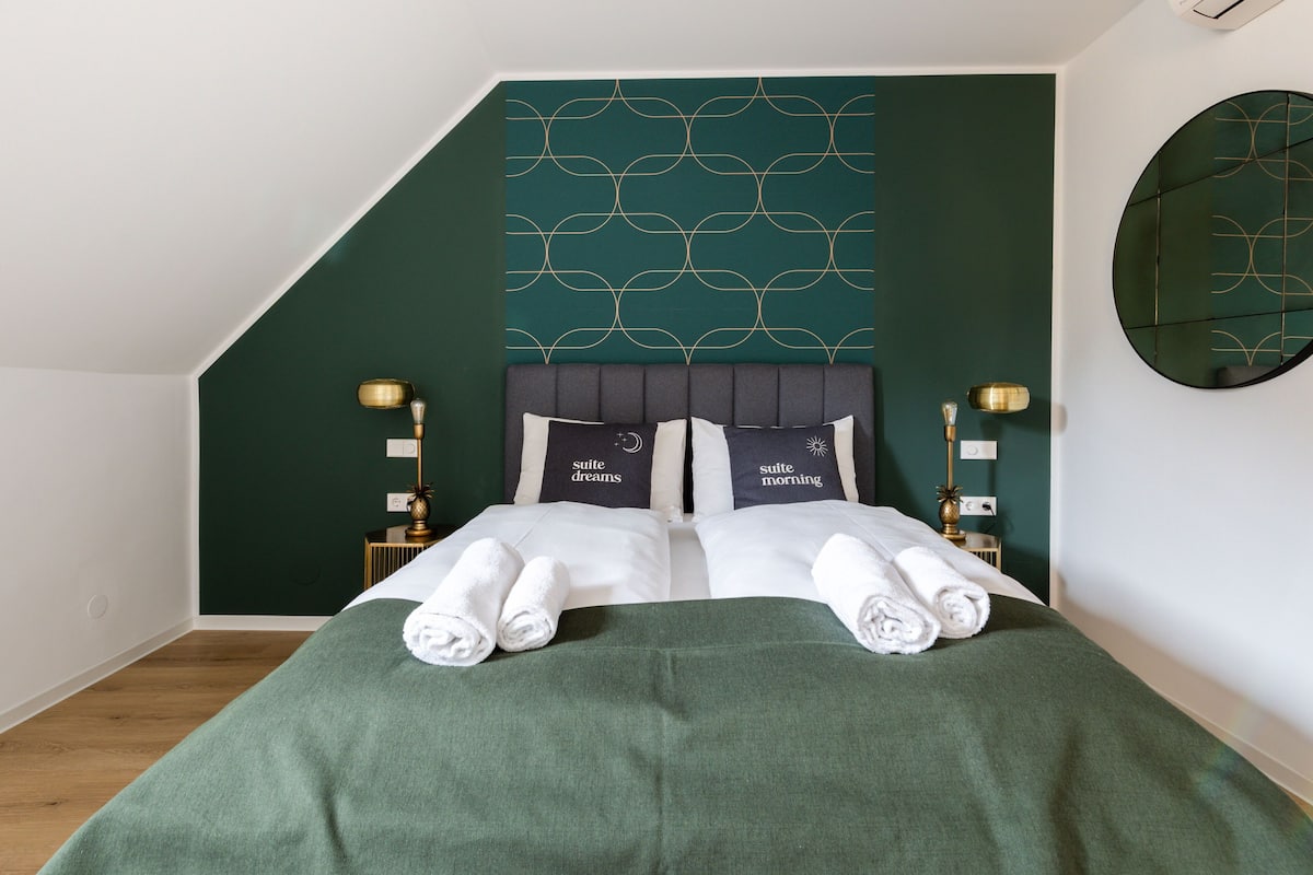 Cama bem arrumada com duas almofadas, um cobre-leito verde e duas toalhas enroladas.