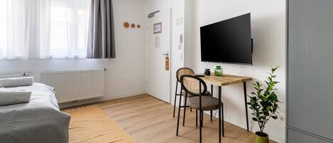 A modern, well-lit room with a bed, a desk, two chairs, a television, and a potted plant.
