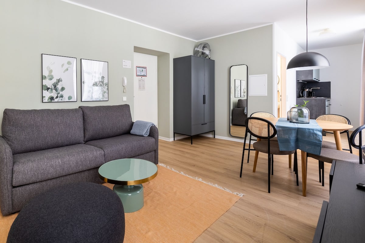 Uma sala de estar moderna com um sofá cinzento, uma mesa de centro redonda e uma área de jantar com mesa e cadeiras de madeira.