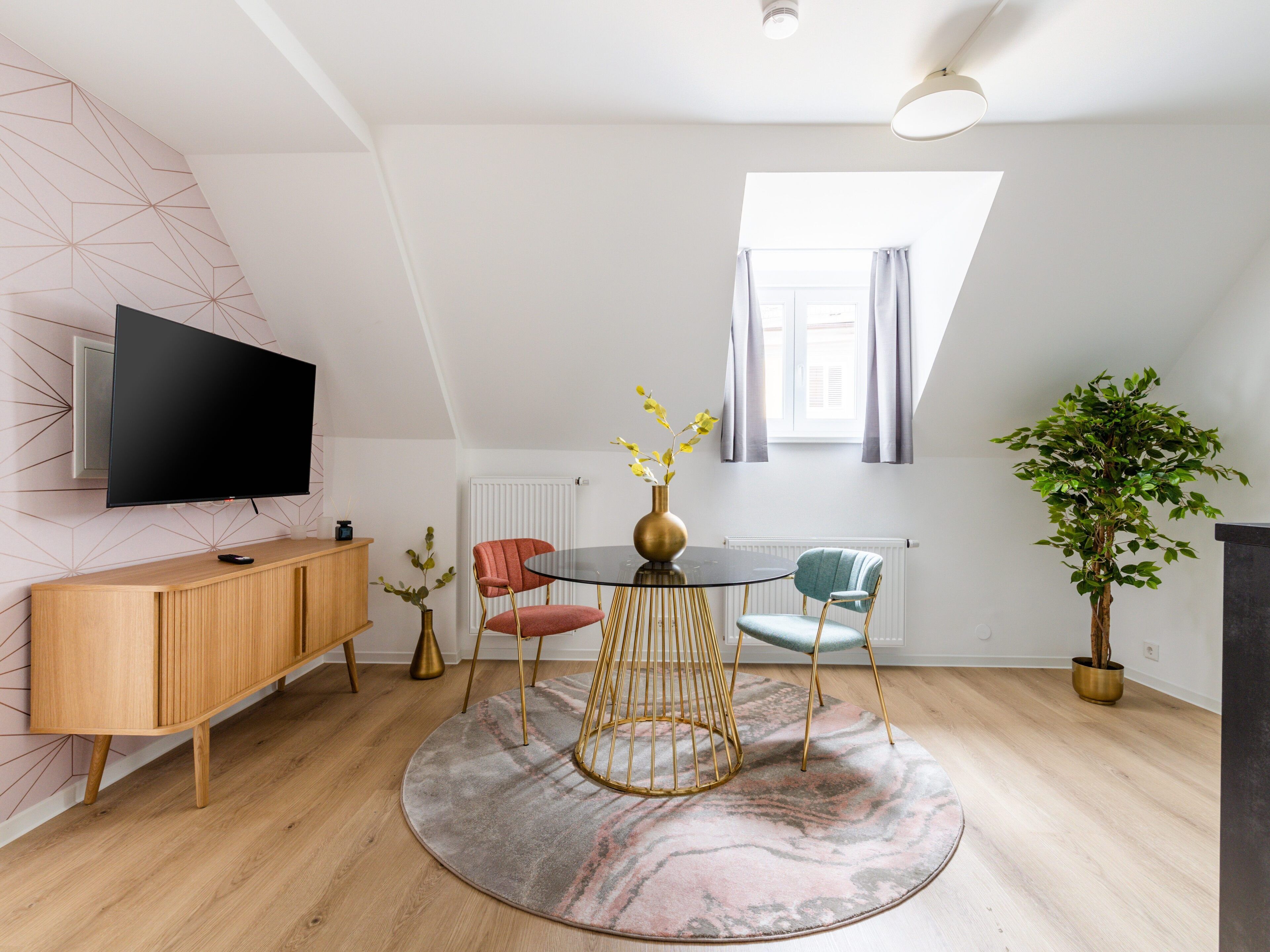 A modern living room with a round glass table, two chairs, a TV, a wooden cabinet, and a potted plant.