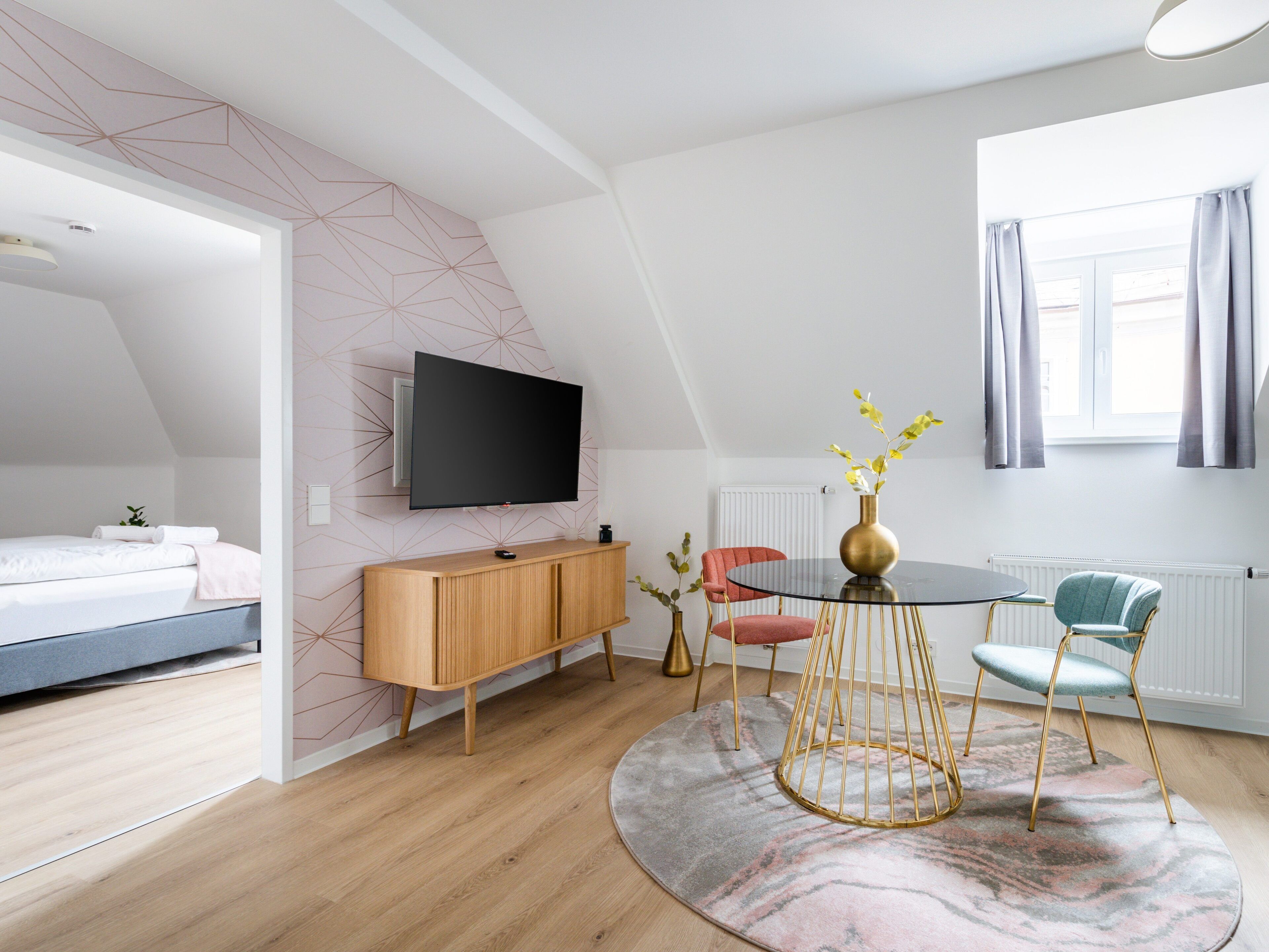 Ett modernt sovrum med en säng, en TV som är monterad på väggen, en byrå i trä, ett runt matbord med stolar och ett fönster med gardiner.
