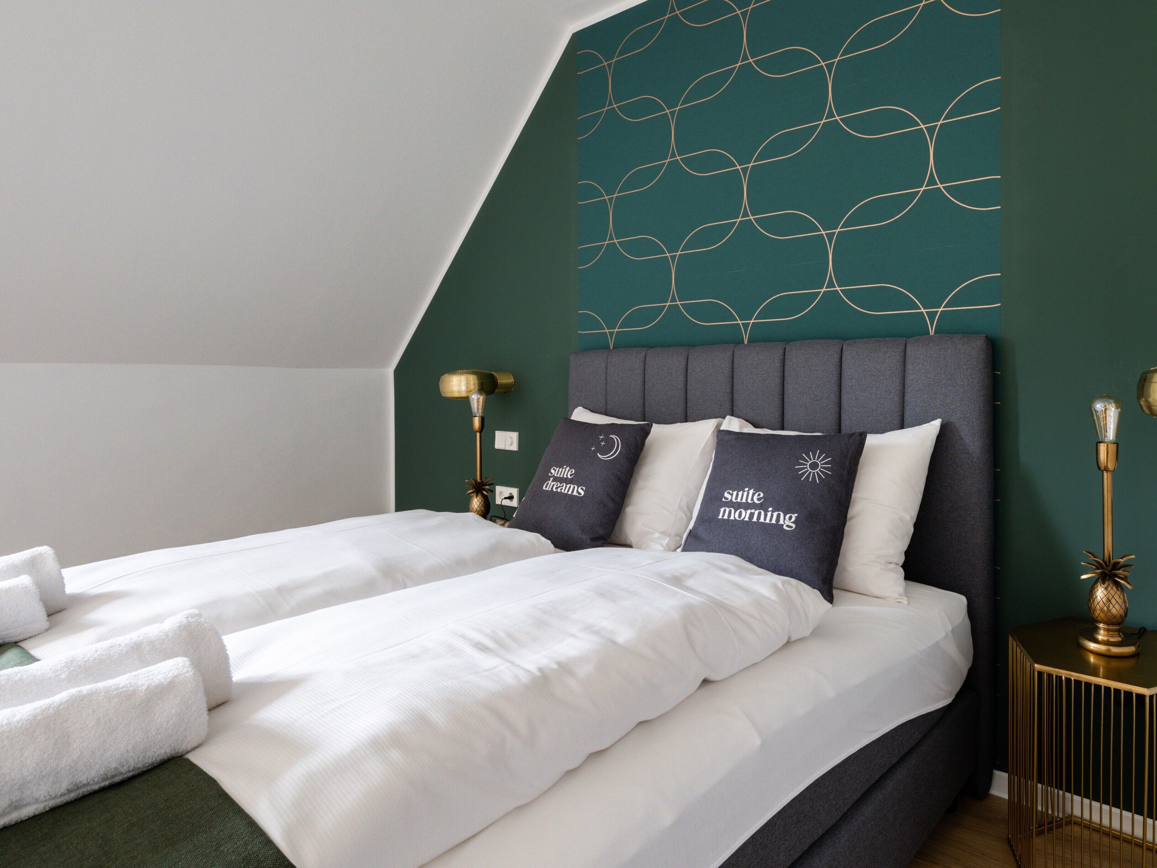A double bed with white linens, two pillows with text, a gold lamp, and a green accent wall.