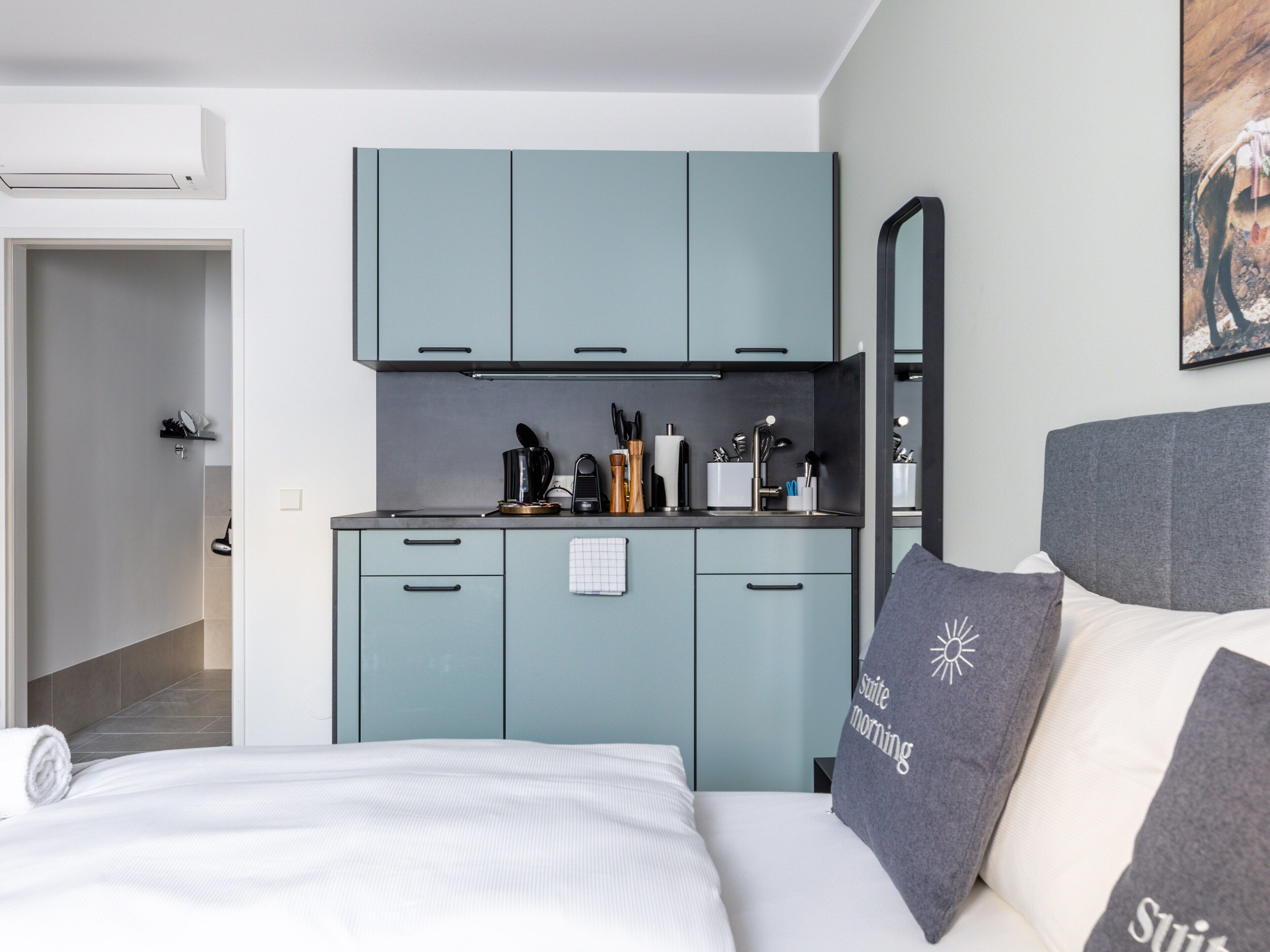 Un dormitorio moderno con cama, mesita de noche, kitchenette con armarios y un baño visible a través de una puerta abierta.