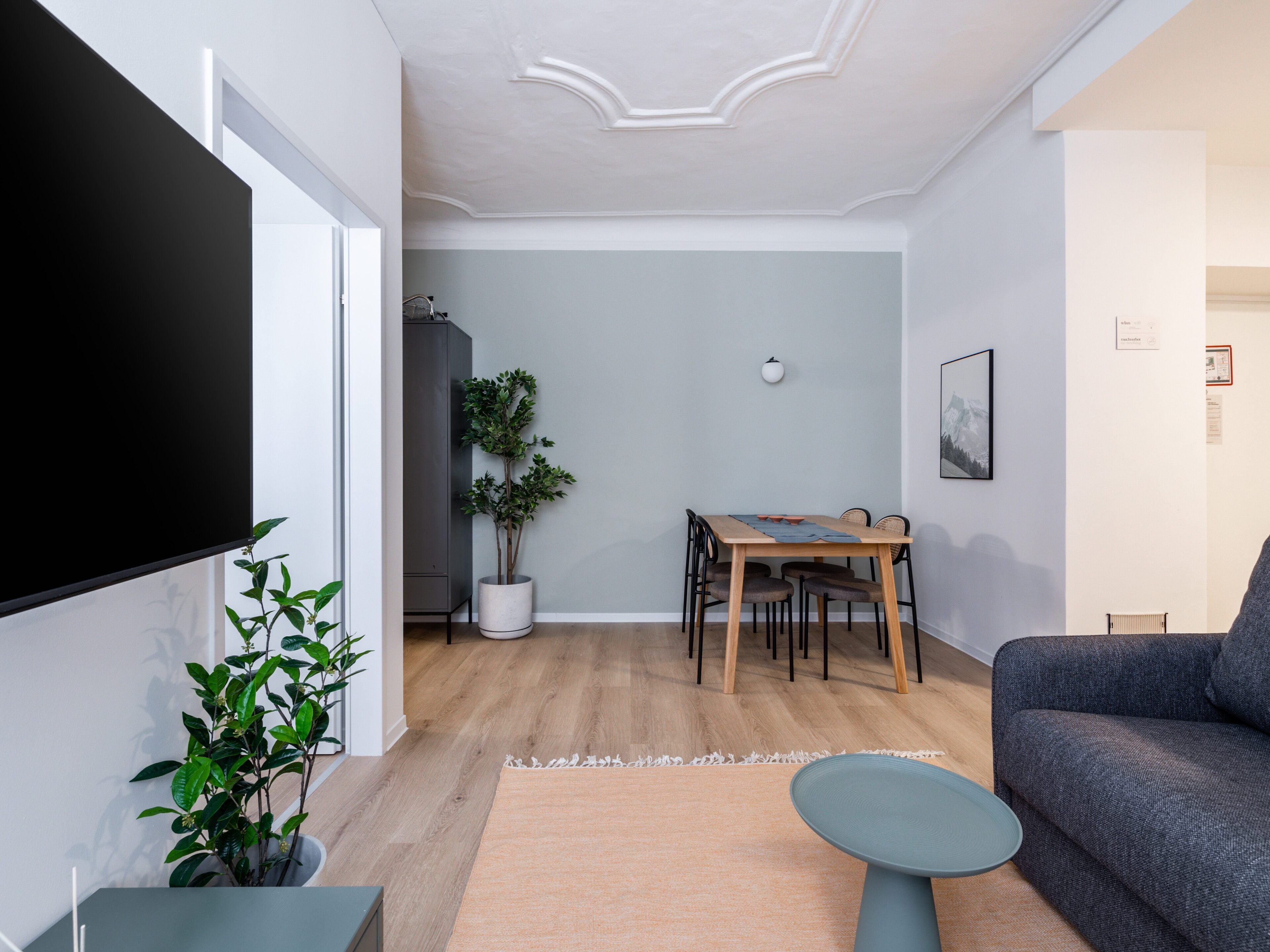 A living room with a flat-screen TV, a grey sofa, a dining table with chairs, a potted plant, and a small round table.