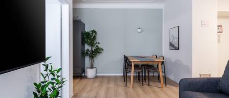 Una sala de estar con una televisión de pantalla plana, un sofá gris, una mesa de comedor con sillas, una planta en maceta y una mesita redonda.