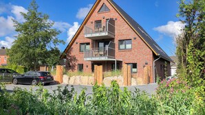 Ein zweigeschossiges Ziegelhaus mit Balkon im zweiten Stock, ein Auto davor und ein Garten mit verschiedenen Pflanzen und Blumen.