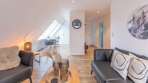 Ein modernes Wohnzimmer mit einer Couch, einem Couchtisch und Dekorationselementen. Im Hintergrund ist ein Essbereich mit Tisch und Stühlen zu sehen.