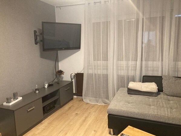 A modern bedroom with a flat-screen TV mounted on the wall, a bedside table with a lamp, and a bed with a grey blanket.