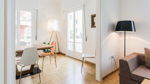 Ein modernes Wohnzimmer mit Holzboden, einer Couch, einem Esstisch mit Stühlen, einer Stehlampe und einem großen Fenster mit Blick auf Gebäude.
